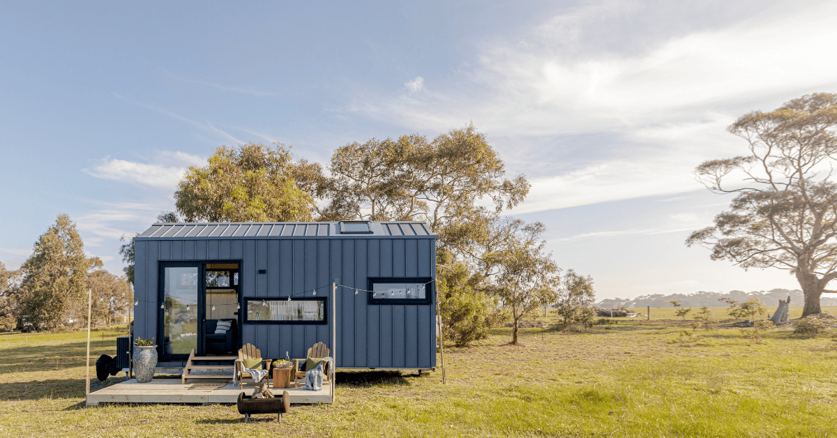 Tiny house on wheels set in a grassy open field with a bright sky and surrounding trees.