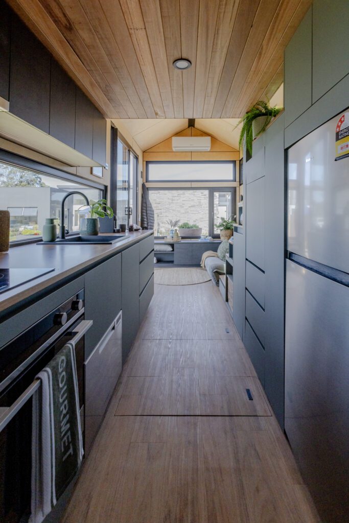 Interior view of a modern tiny home  on wheels kitchen with sleek cabinetry, integrated appliances, and large windows providing natural light, leading to a cozy living area in the background.