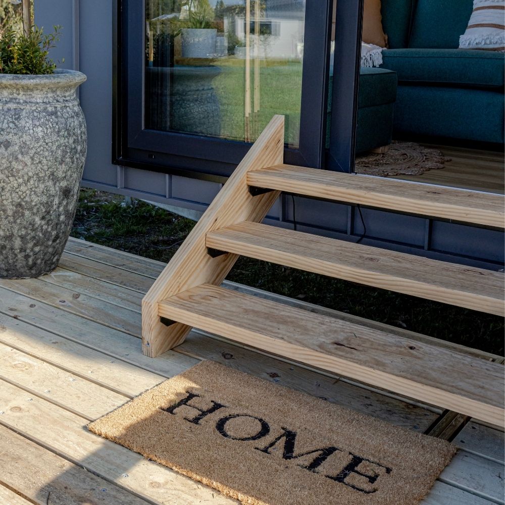 Sturdy treated pine steps placed on a modular deck outside a tiny home entrance, featuring a welcoming doormat and potted plant.